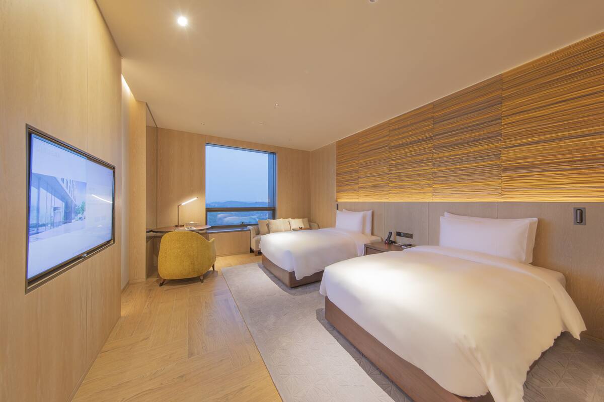 A modern hotel room with two beds, a large window, a view of the city, and a wooden wall panel.