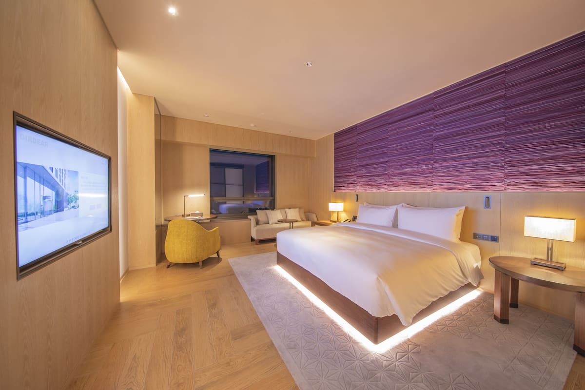 A modern hotel room with a large bed, a sofa, a desk, and a television mounted on the wall.