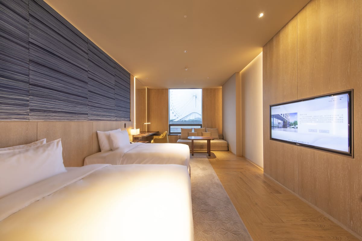 A modern hotel room with two beds, a desk, a chair, and a large TV screen.