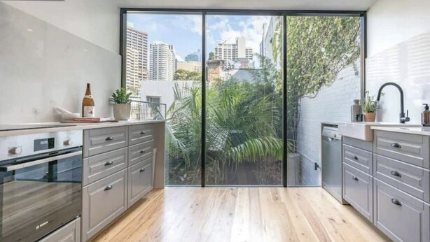 A modern kitchen with grey cabinets, a built-in oven, and a large sliding glass door leading to a balcony with a city view. - A Touch of Soho: Luxurious Historic Residence (Darlinghurst)