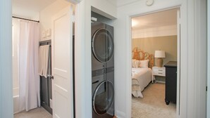 A laundry room with two dryers, a shower area, and a bedroom with a bed, nightstand, and lamp.