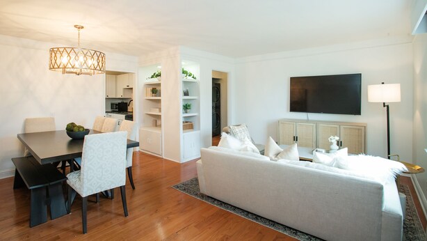 A modern living room with a sofa, dining table, and chairs. There is a kitchen area in the background with cabinets and a TV mounted on the wall.