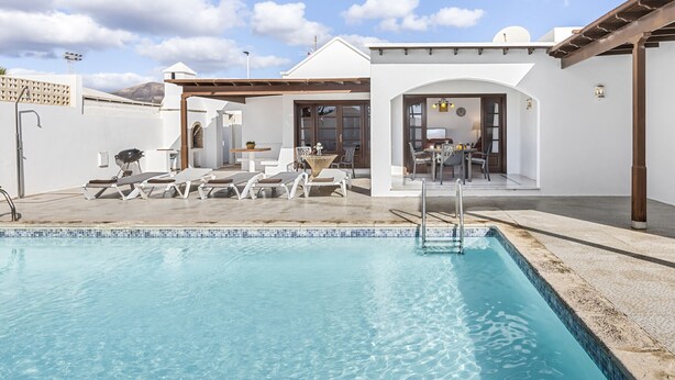 A swimming pool with lounge chairs, a covered patio area, and a view of the interior of a building with a dining area. - Villa Eva - Villasexperience (Puerto del Carmen)