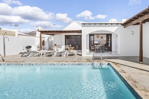 A swimming pool with lounge chairs, a covered patio area, and a view of the interior of a building with a dining area. - Villa Eva - Villasexperience (Puerto del Carmen)