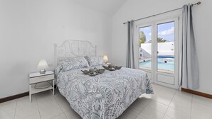A bedroom with a bed, nightstand, lamp, and a view of a swimming pool through a glass door. - Villa Eva - Villasexperience (Puerto del Carmen)