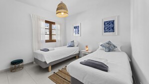 A bedroom with two beds, a bedside table with a lamp, and a window with curtains. - Casa Estay | Holiday Apartment in Lanzarote - Spain (Playa Honda)