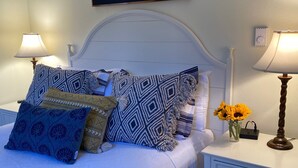 Ein ordentlich bezogenes Bett mit gemusterten Kissen, zwei Nachttischlampen, ein kleiner Tisch mit einer Vase voller Sonnenblumen und ein Thermostat an der Wand.