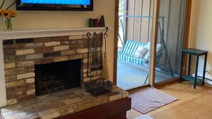 Ein Wohnzimmer mit Kamin aus Backstein, einem flachbildfernseher darüber und einer Glastür, die zu einem Außenbereich mit Terrasse und einem Stuhl führt.