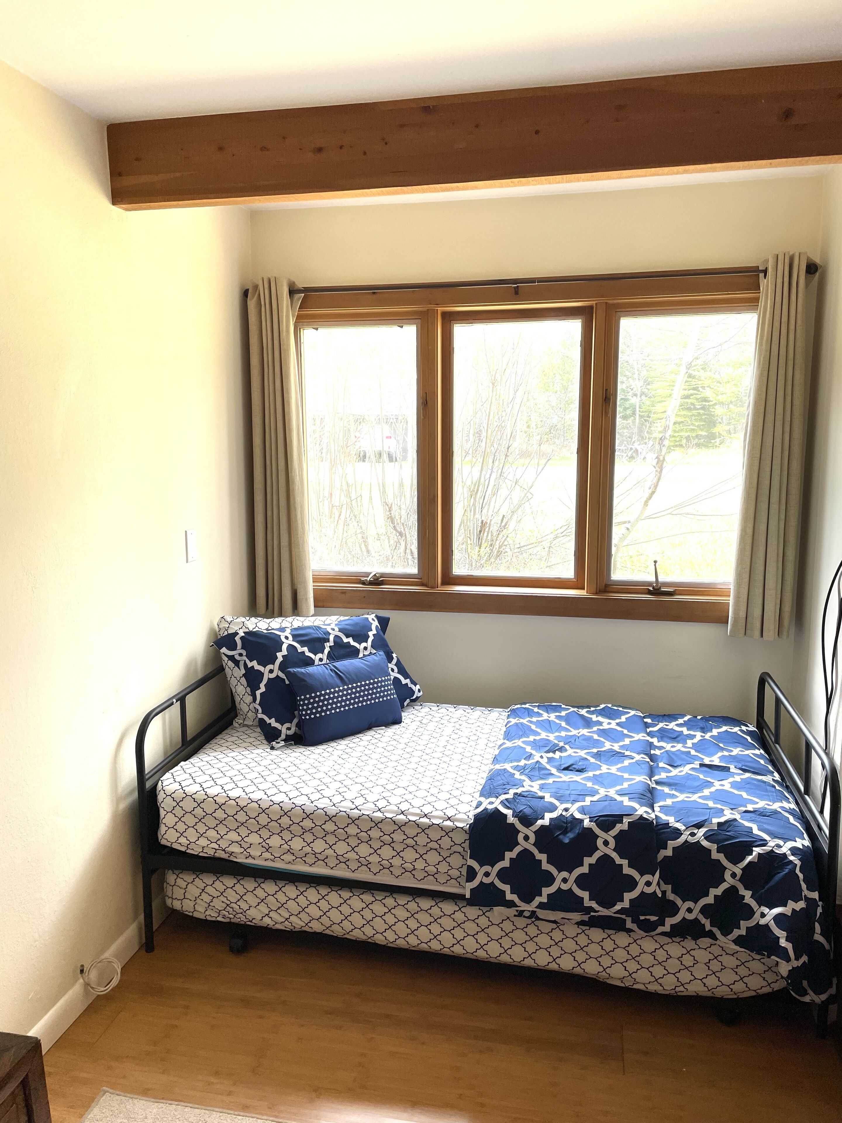 A small, single-bed room with a wooden ceiling, a window with curtains, and a patterned bedspread.