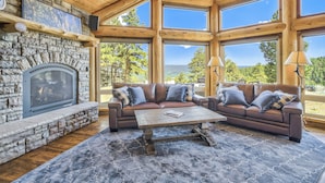 Un salon chaleureux avec une cheminée en pierre, un plafond en bois et de grandes fenêtres offrant une vue panoramique.