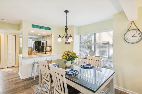 A dining area with a glass table, white chairs, a chandelier, and a clock on the wall.