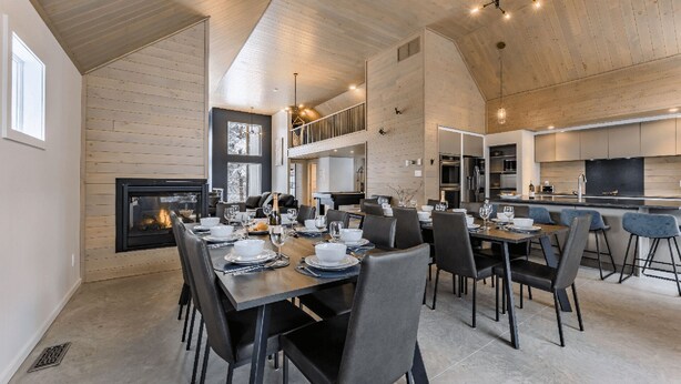 A modern dining area with a fireplace, a kitchen in the background, and a loft space above.