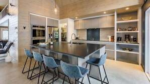 A modern kitchen with a central island, bar stools, stainless steel appliances, and a wooden ceiling.