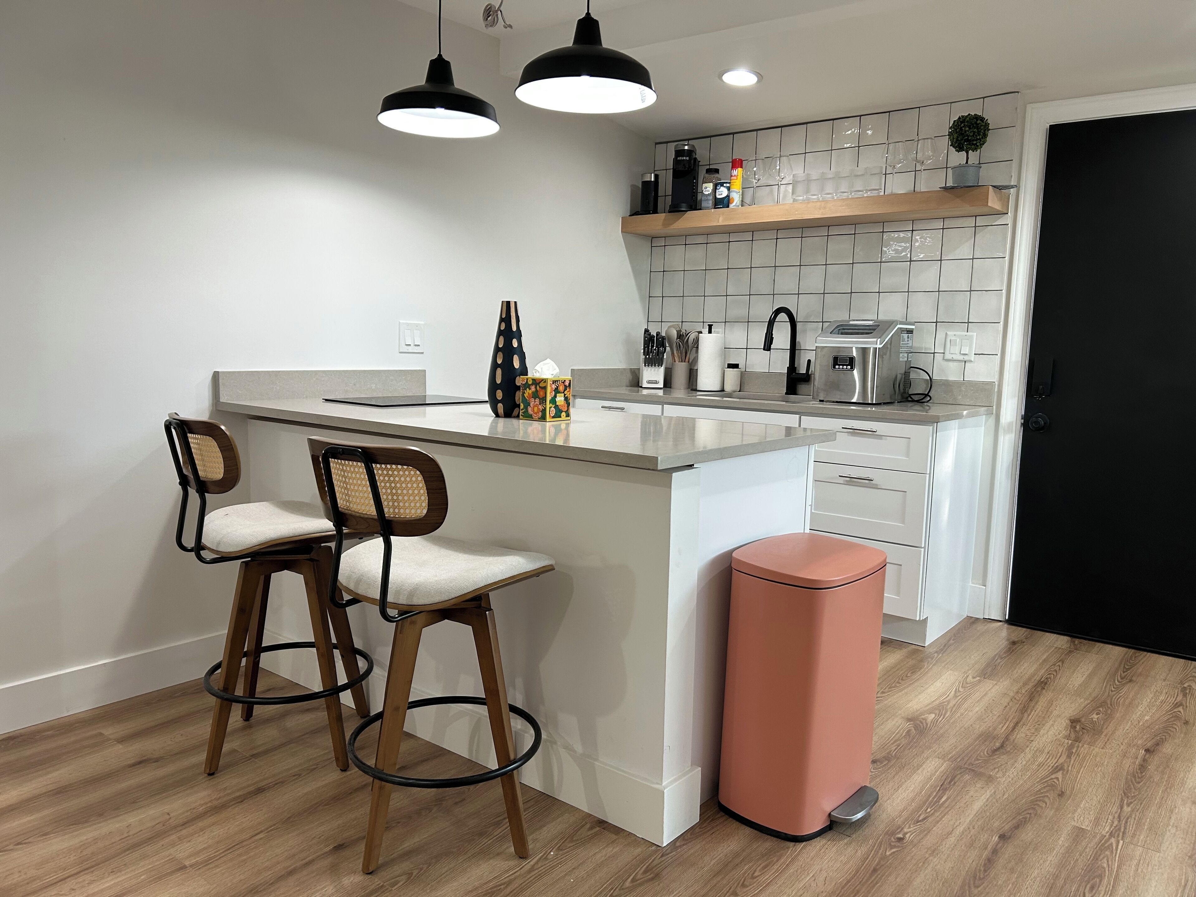 A modern kitchen with a central island, two bar stools, a trash bin, and a black door.