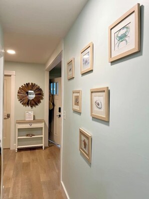 A hallway with framed artwork on the walls, a wooden console table with a mirror, and a door leading to another room.