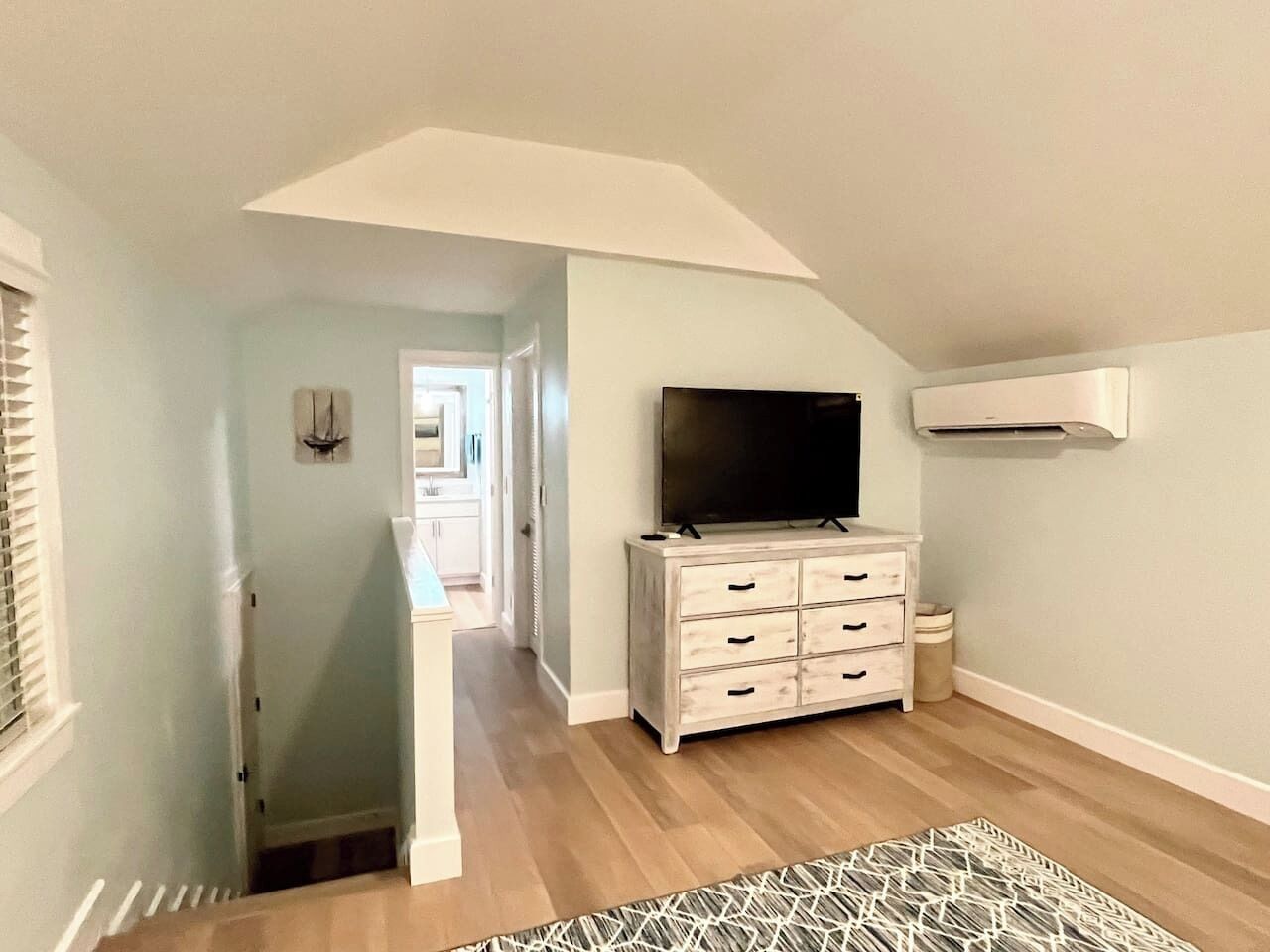 A room with a flat-screen TV on a dresser, a patterned rug, and a hallway leading to another room.
