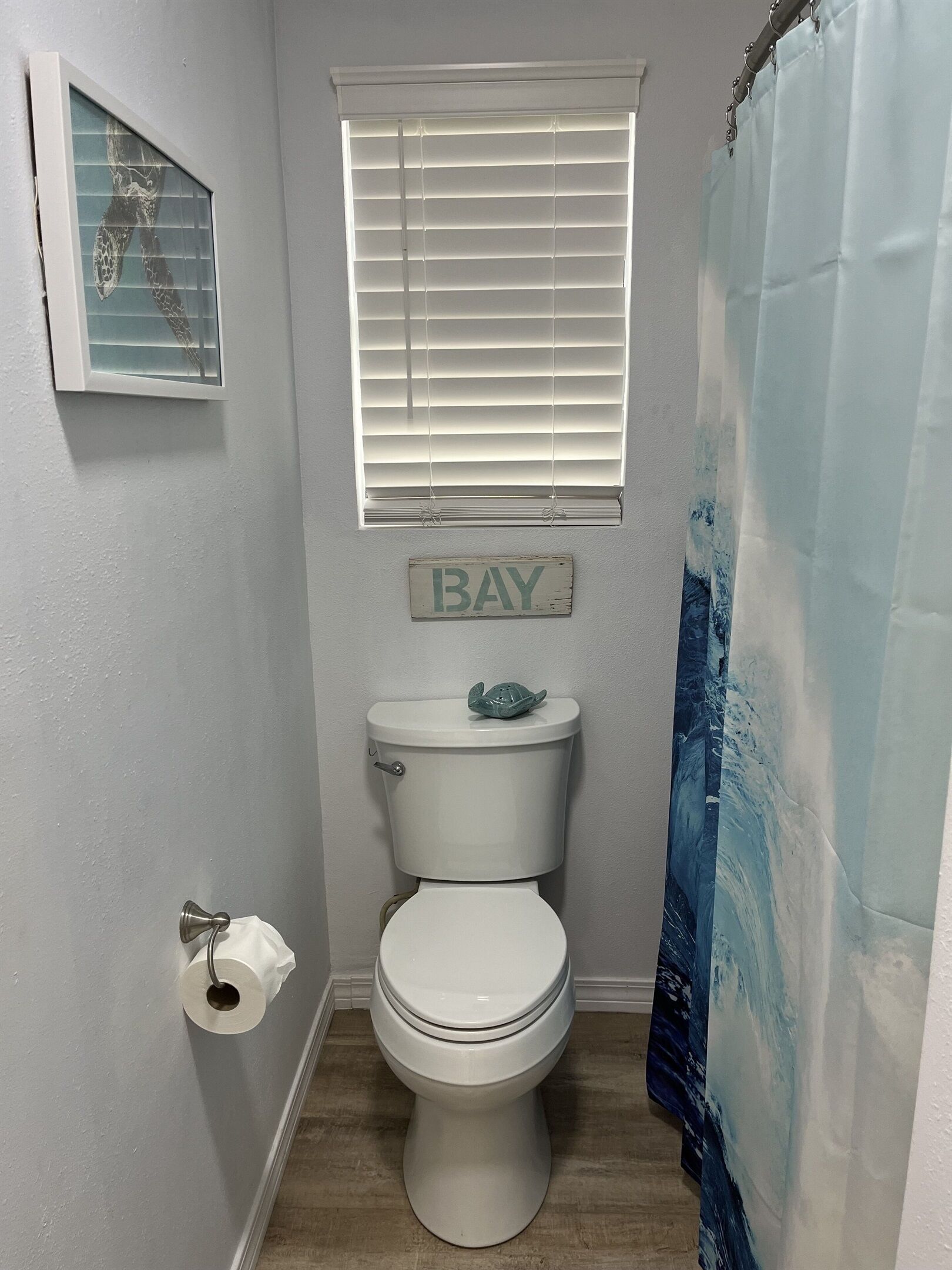 A small bathroom with a toilet, a window with blinds, a 'BAY' sign, and a shower curtain.