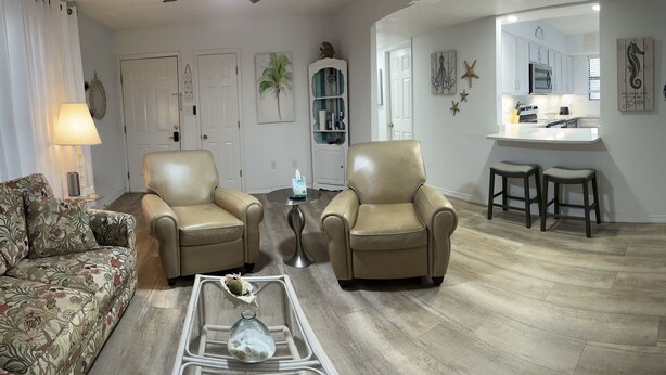A living room with two beige armchairs, a floral sofa, a glass coffee table, and a kitchen area with white cabinets and a microwave. - B2 Boujee, a ground floor, cozy condo, just a short walk to the beach.. (Gulf Shores)