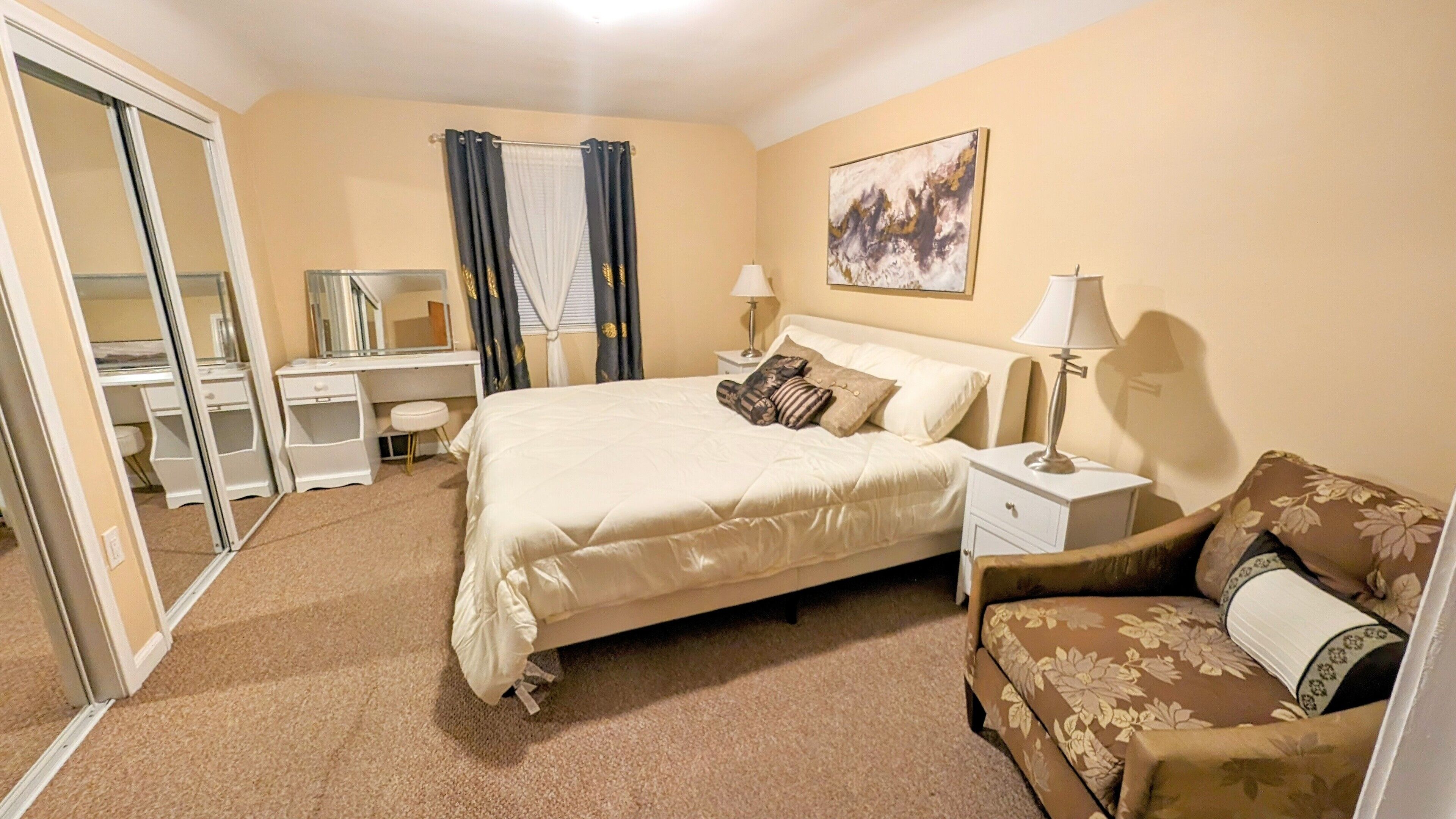 A hotel room with a bed, a desk, a chair, a nightstand, and a painting on the wall.
