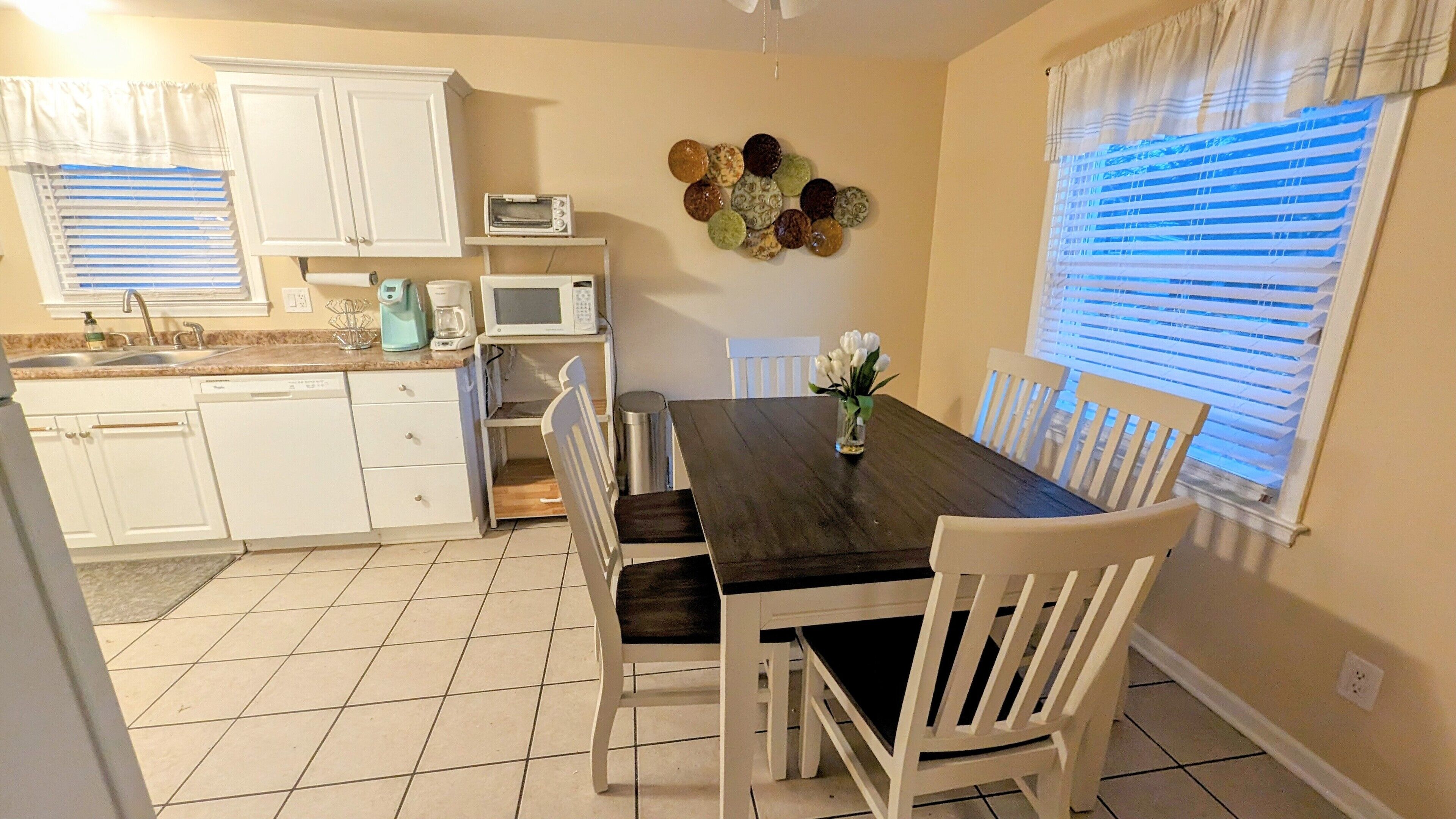 A dining area with a dark wooden table, white chairs, a vase with flowers, a microwave, and a wall-mounted clock.