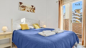 Ein Hotelzimmer mit Bett, Nachttisch, Lampe, einem Gemälde an der Wand und Blick auf Gebäude durch das Fenster.