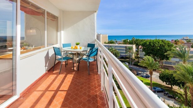 A balcony with a table and chairs, overlooking a cityscape and the sea.