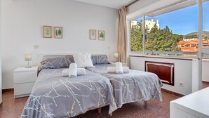 A hotel room with two beds, a large window with a view of buildings and trees, and a small desk.