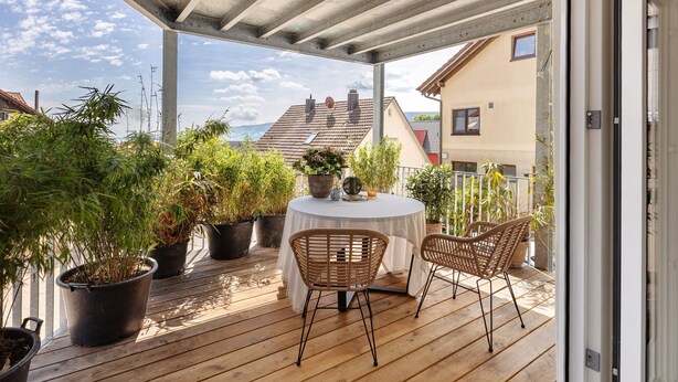 Una terraza de madera con una mesa redonda y dos sillas de mimbre, rodeada de macetas con plantas.