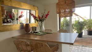 Un comedor con mesa de madera, sillas de mimbre, un florero con flores, una lámpara colgante de fibra natural y vistas al exterior con vegetación.