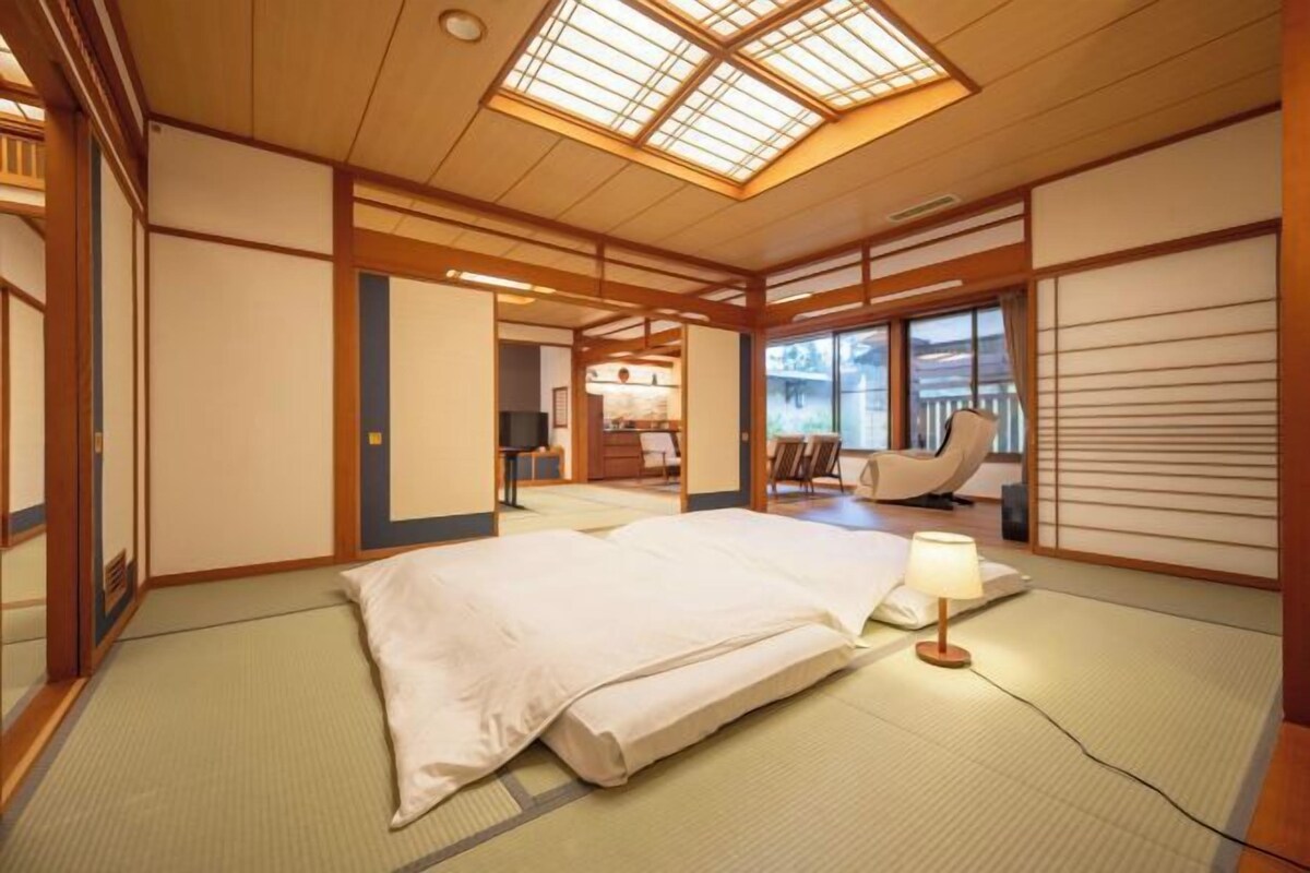 Una habitación tradicional japonesa con suelo de tatami, una cama con edredón blanco, un escritorio con lámpara y vistas al exterior a través de amplias ventanas.