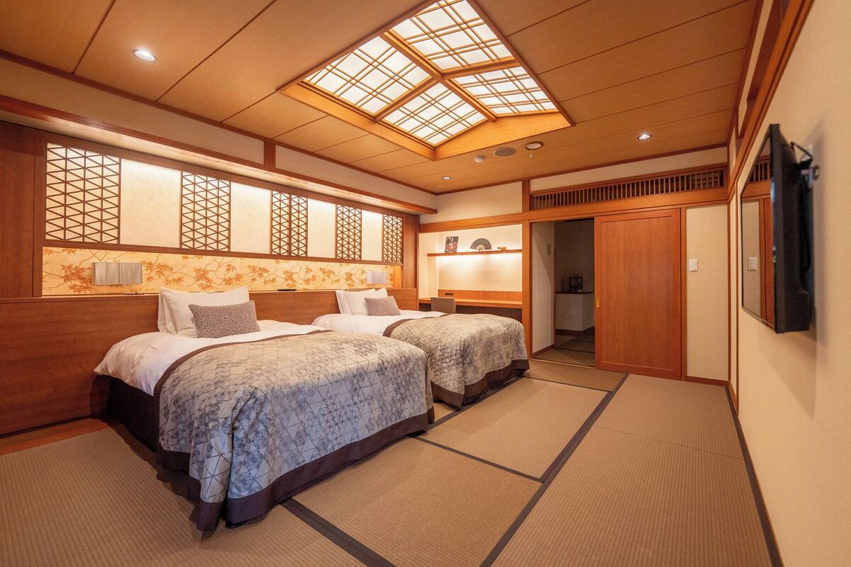 A traditional Japanese-style hotel room with tatami flooring, two beds with patterned bedding, a skylight, and a sliding door leading to a bathroom.