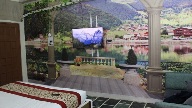 Habitación con un televisor grande mostrando un paisaje pintoresco, una cama con colcha estampada floral y vista a un lago con montañas al fondo.