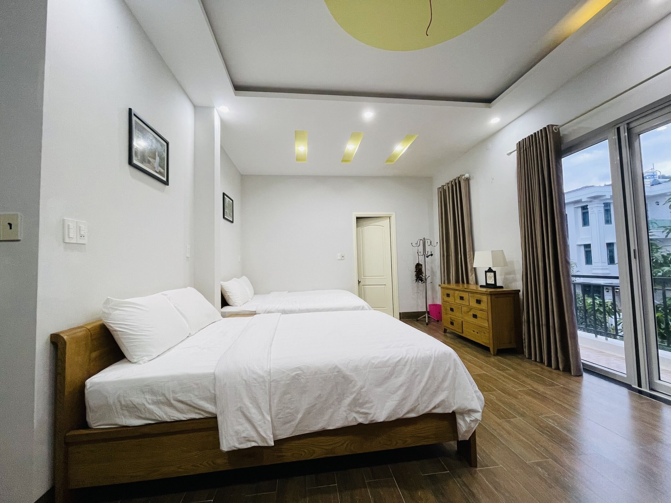 A modern hotel room with two beds, a wooden dresser, a coat rack, and a large window with a view of buildings.