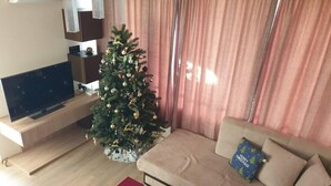 A living room with a Christmas tree, a beige sofa, a red coffee table, and a flat-screen TV on a wooden stand.