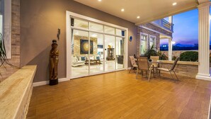 Un patio amplio con suelo de madera, un comedor con sillas y una mesa, y vista del interior a través de puertas de cristal.
