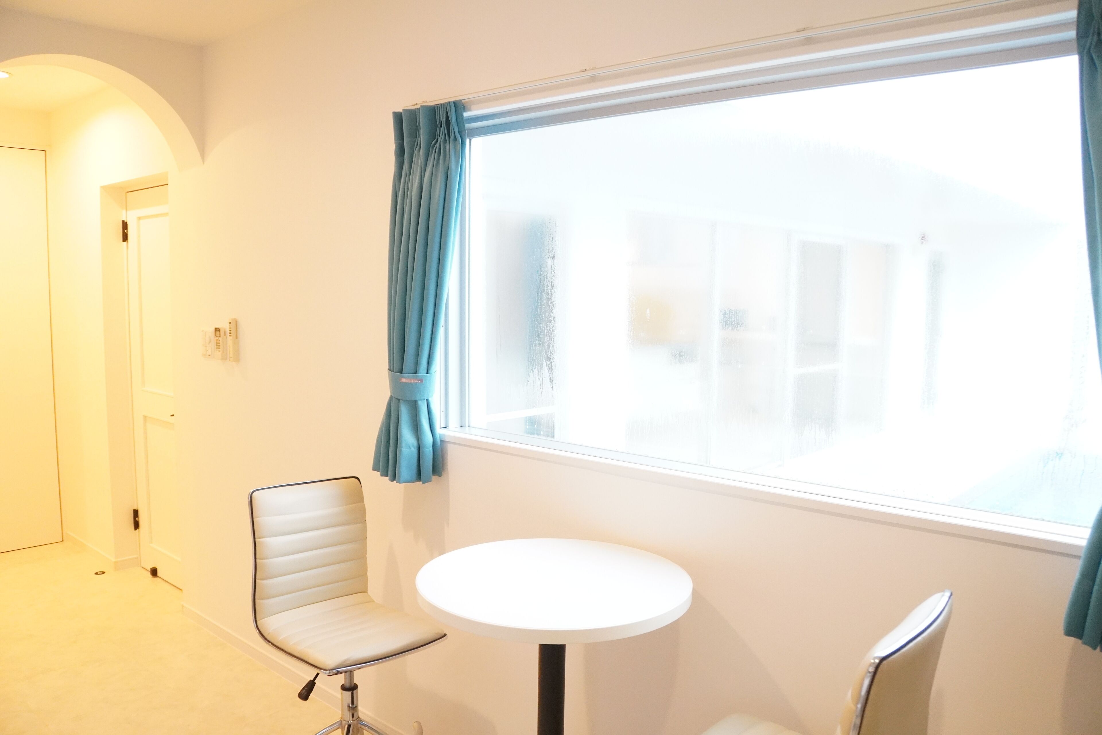 A room with a round table and two chairs, a door, and a window with curtains.