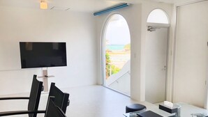Una habitación moderna con televisión de pantalla plana, una mesa de comedor con sillas, un pequeño rincón de descanso y un balcón con vistas a la playa.