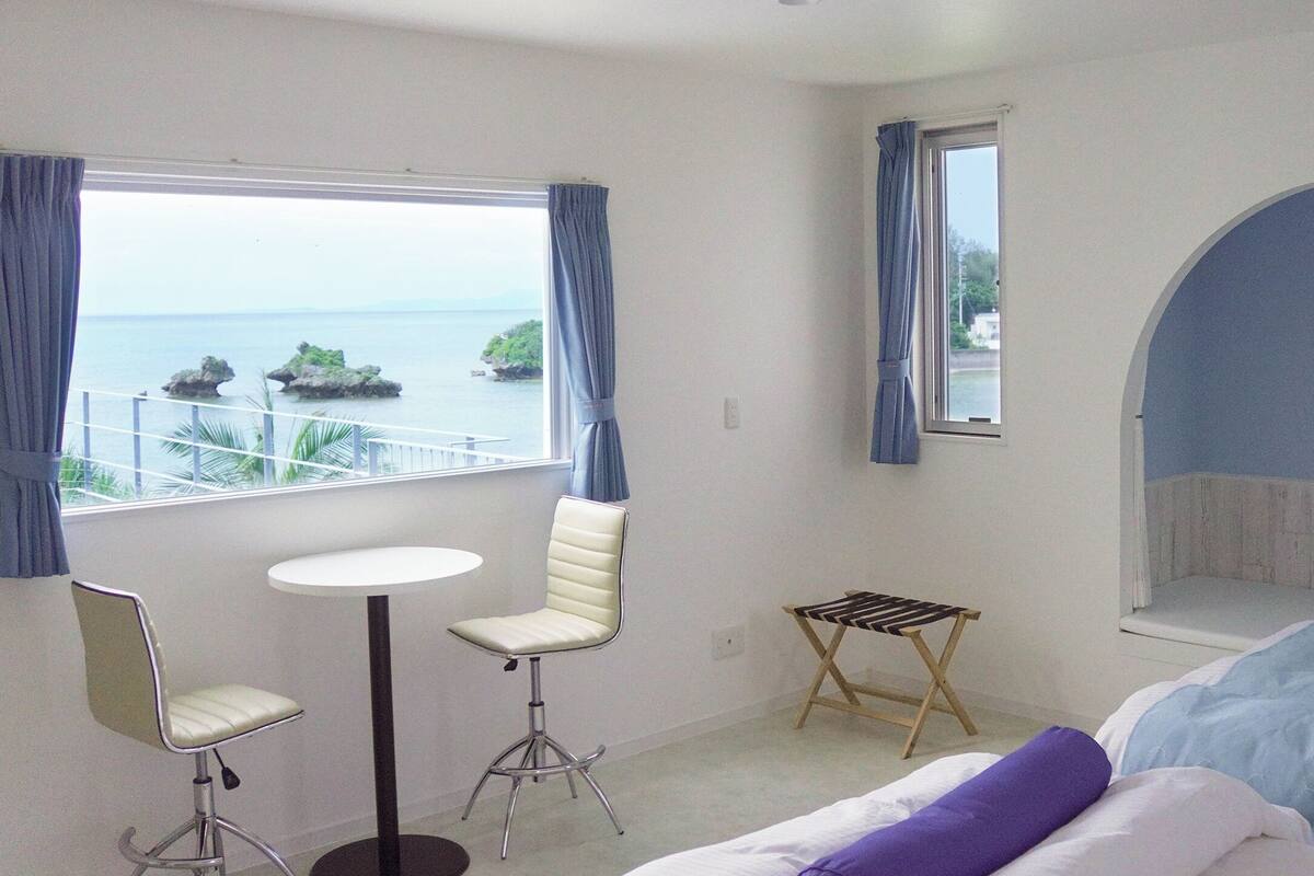 A hotel room with a bed, a small table, two chairs, a window with a view of the sea, and a unique arched ceiling.