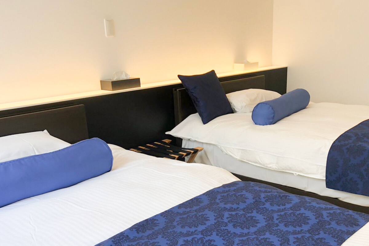 Two single beds with white and blue bedding, a bedside table with a lamp, and a small box on the wall.