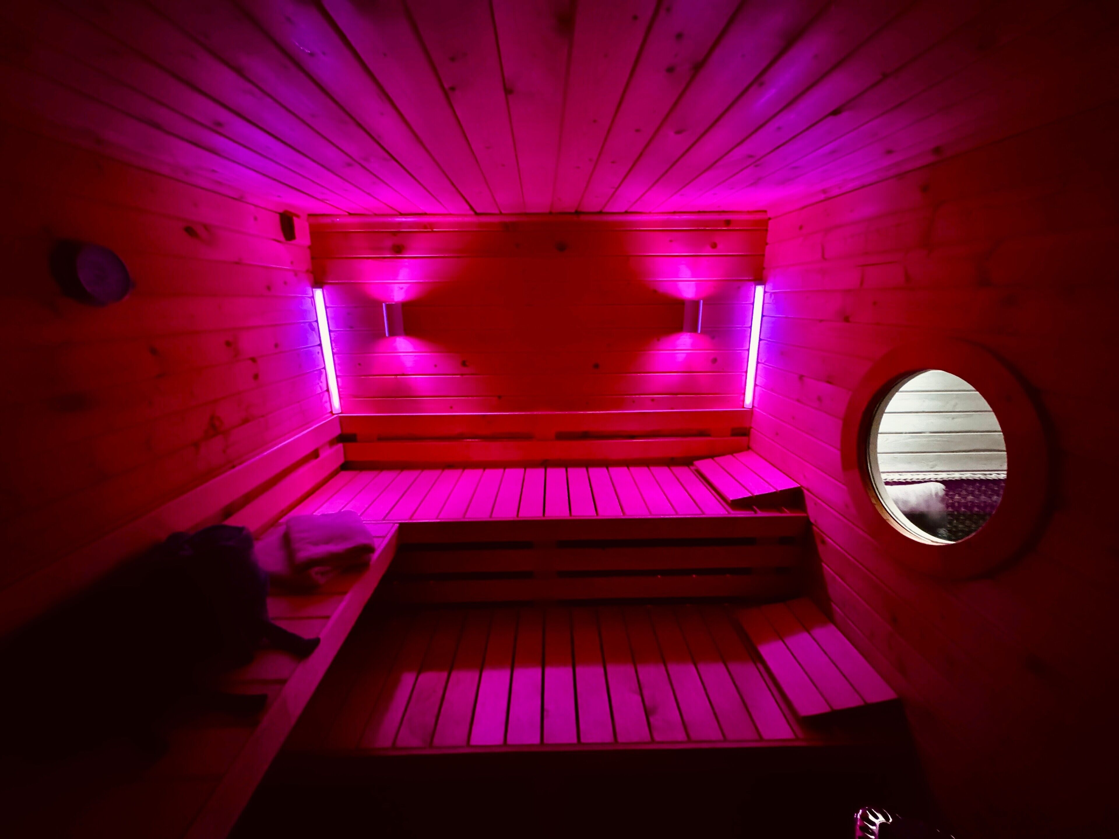 Un sauna avec des bancs en bois, une fenêtre circulaire et un éclairage rose.