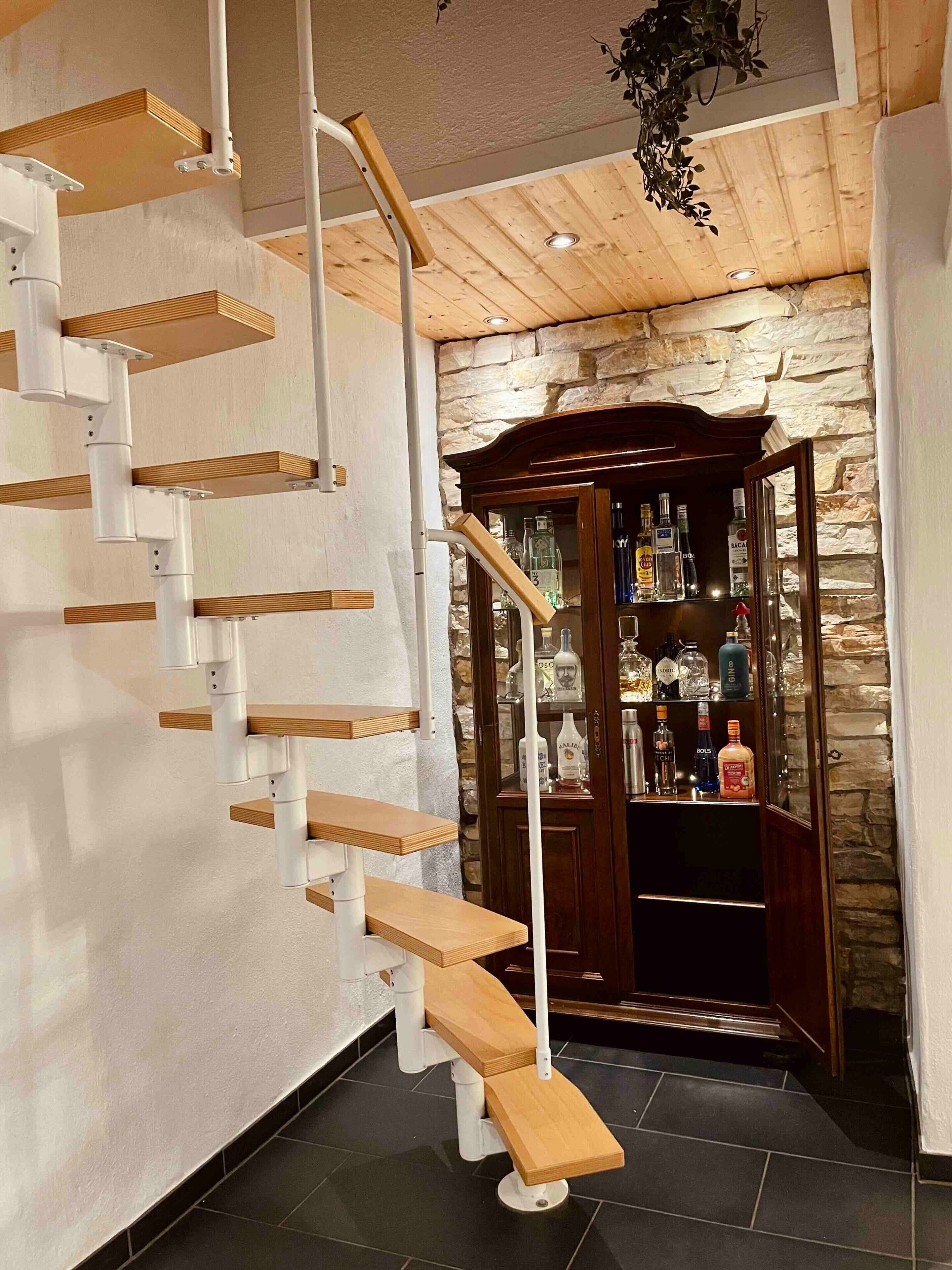 A spiral staircase with wooden steps and white railings, leading to a stone wall with a liquor cabinet.