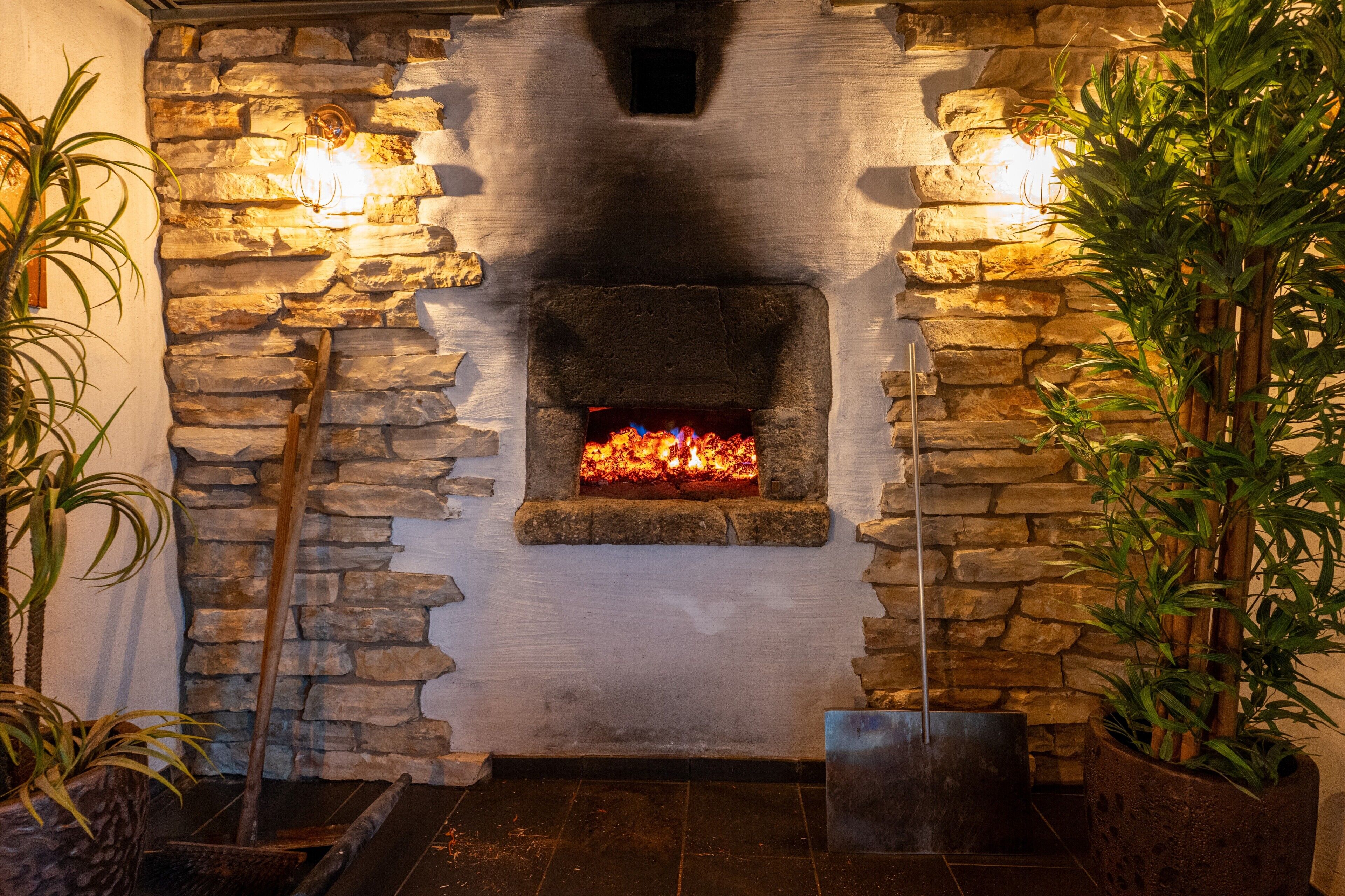 A stone fireplace with a fire, flanked by two wall-mounted lights, and surrounded by potted plants.