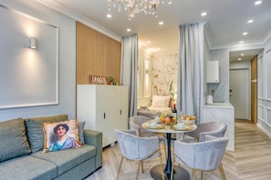 A modern living room with a sofa, a dining table with chairs, a chandelier, and a kitchen area in the background.
