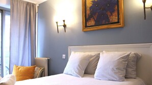 Un dormitorio con cama, almohadas blancas, radiador, ventana con cortinas y un cuadro en la pared.