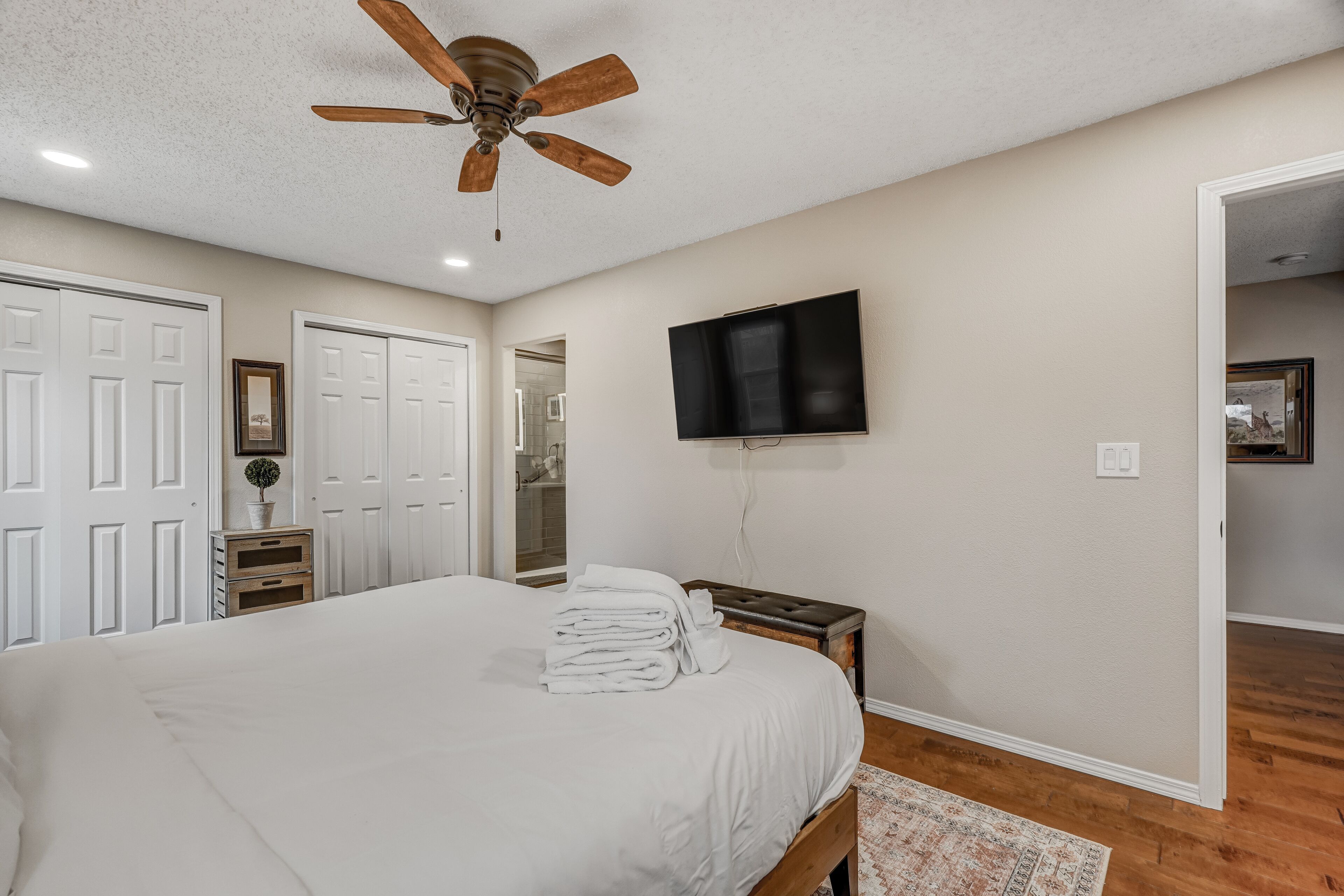 A bedroom with a large bed, a ceiling fan, a TV mounted on the wall, and a wooden floor.