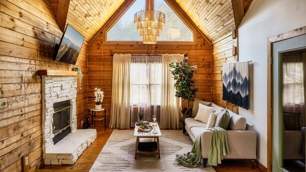 A cozy log cabin interior with a stone fireplace, a sofa, a coffee table, and a large window with curtains. - New Hot Tub, Hammocks, Pet Friendly, Romantic Honeymoon Mountain Getaway (Pigeon Forge)