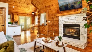 A cozy cabin interior with a stone fireplace, wooden walls, a dining area, and a TV mounted above the fireplace. - New Hot Tub, Hammocks, Pet Friendly, Romantic Honeymoon Mountain Getaway (Pigeon Forge)