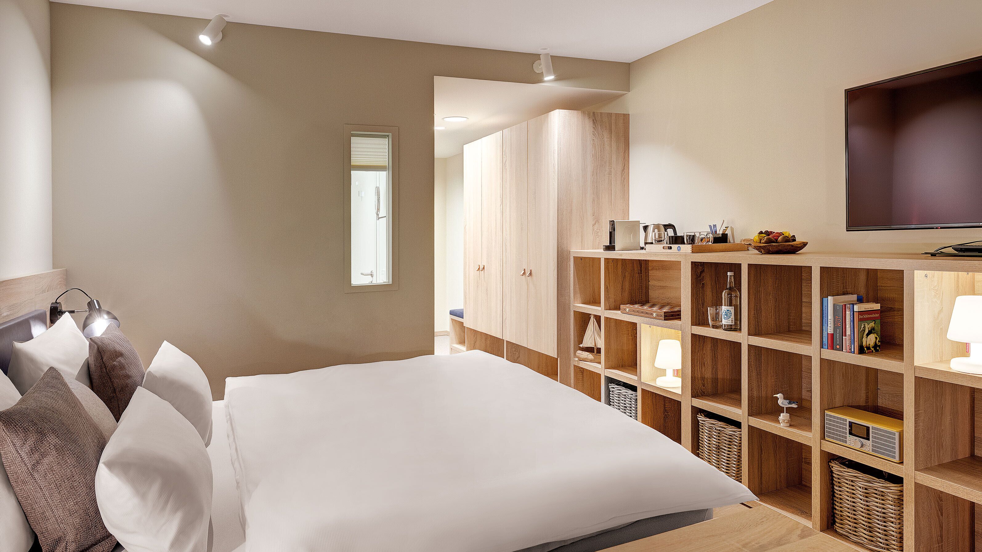 A bedroom with a large bed, wooden wardrobe, shelves with books and a radio, and a TV on the wall.