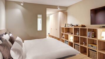 A bedroom with a large bed, wooden wardrobe, shelves with books and a radio, and a TV on the wall.