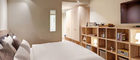 A bedroom with a large bed, wooden wardrobe, shelves with books and a radio, and a TV on the wall.